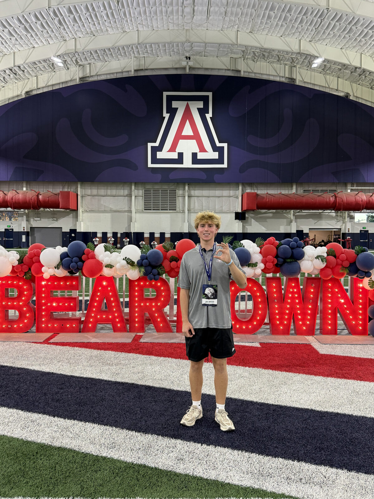 Jax Snyder in front of the lit Beardown marquee sign inside the University of Arizona indoor practice facility, wearing an Arizona polo and his Arizona Football visit credential